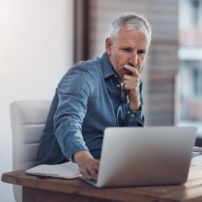 Pensive businessman sitting at desk using a laptop in a modern office.